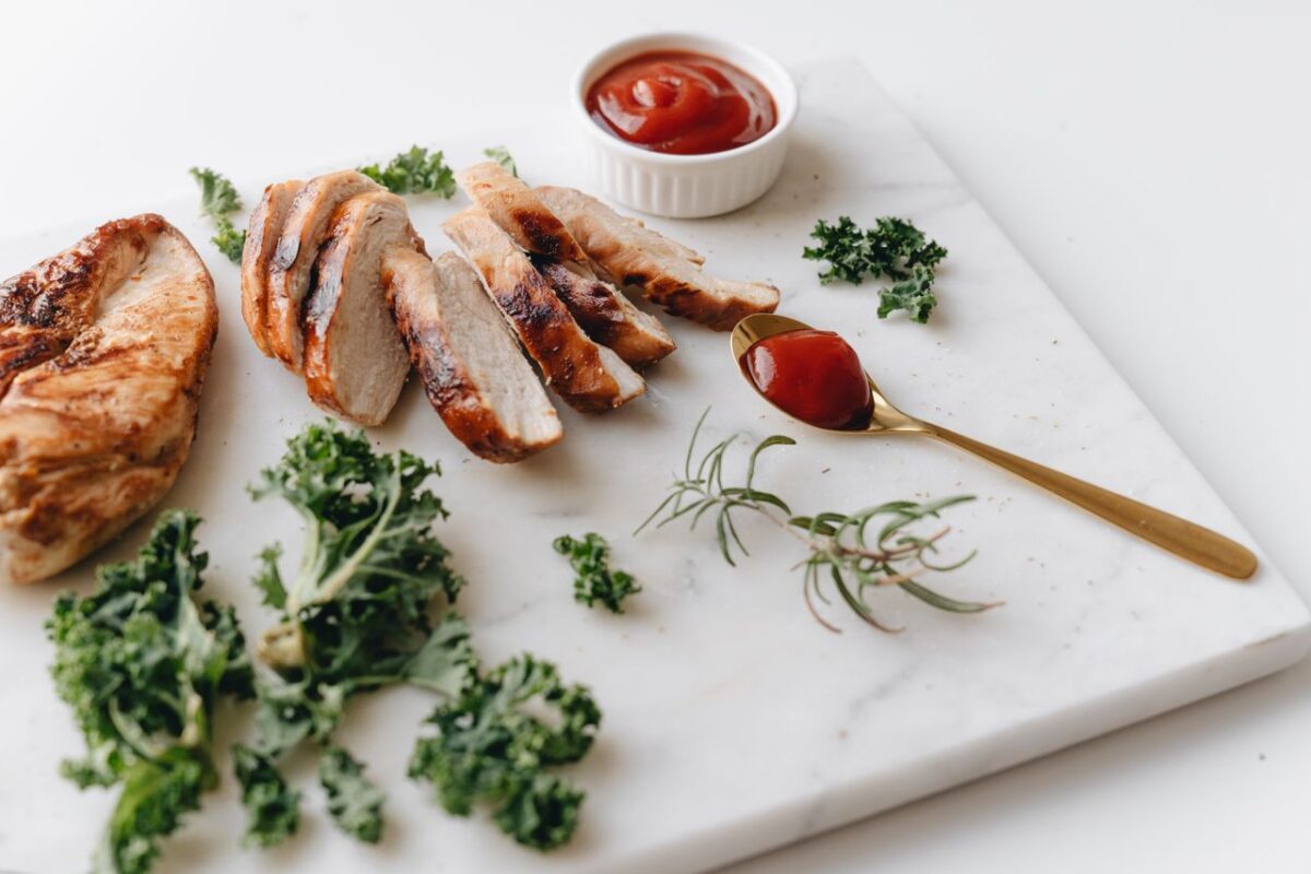 impact-img Sliced grilled chicken with greens and ketchup on a marble board, perfect for a healthy meal.