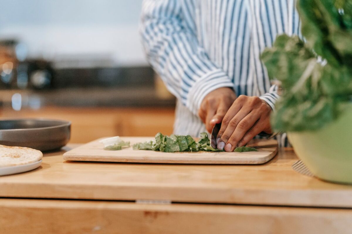 hero-homepage Crop anonymous ethnic male in striped shirt cutting fresh spinach on chopping board while cooking at home